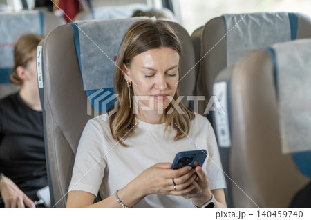 A young woman sits in a train carriage, focused on her smartphone while traveling during the day, surrounded by fellow passengers. 140459740