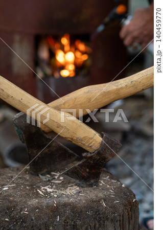 Two wooden handles cross over a black axe blade resting on a wooden stump, surrounded by shavings and a warm campfire glow in the background. Two wooden handles cross over a black axe blade resting on a wooden stump, surrounded by shavings and a warm campfire glow in the background. 140459770