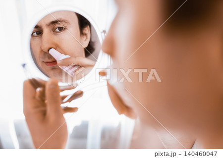 Man looking in a mirror using an electric trimmer for nose hair removal. A daily male grooming routine and personal facial care for health and hygiene. Man looking in a mirror using an electric trimmer for nose hair removal. A daily male grooming routine and personal facial care for health and hygiene. 140460047