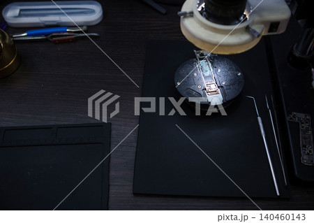 A technician is working on a small electronic device using tools on a dark work surface in a workshop during the afternoon. A technician is working on a small electronic device using tools on a dark work surface in a workshop during the afternoon. 140460143