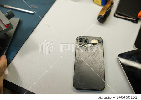 A person examines a cracked smartphone on a work table in a repair shop with various tools and devices present. A person examines a cracked smartphone on a work table in a repair shop with various tools and devices present. 140460152