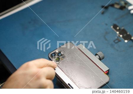 Person removes parts from a smartphone in a workshop. The process involves careful disassembly using tools on a blue surface. 140460159