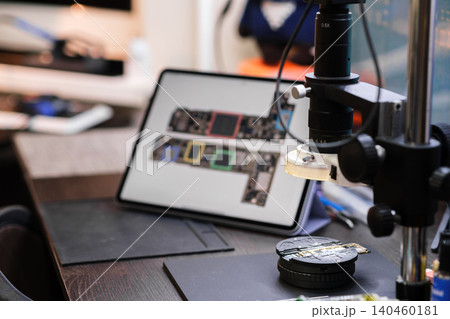 A magnifying device is set up on a workbench next to a tablet displaying circuit board designs. Tools are arranged nearby for electronics work. A magnifying device is set up on a workbench next to a tablet displaying circuit board designs. Tools are arranged nearby for electronics work. 140460181