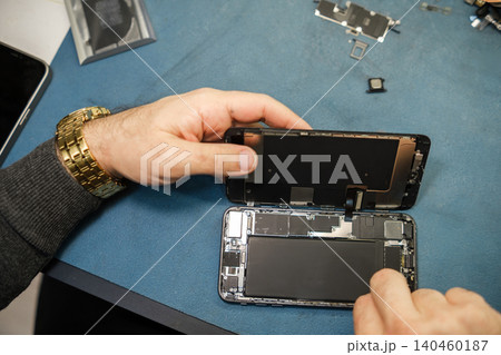 A technician works on a smartphone, carefully removing the back cover while tools and components are organized on a blue surface. A technician works on a smartphone, carefully removing the back cover while tools and components are organized on a blue surface. 140460187