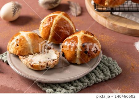 Freshly baked and glazed hot cross buns arranged on a grey plate with one sliced and buttered, featuring a rustic Easter background with feathers and eggs 140460602