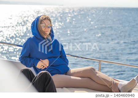 A person sits comfortably on a yacht deck, dressed in a blue hoodie and reflective sunglasses, relaxing on a sunny day by the ocean with gentle waves. A person sits comfortably on a yacht deck, dressed in a blue hoodie and reflective sunglasses, relaxing on a sunny day by the ocean with gentle waves. 140460940