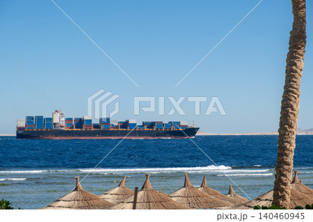 A large container ship glides over the blue sea, with palm trees and umbrellas on the sandy beach, creating a peaceful coastal scene. A large container ship glides over the blue sea, with palm trees and umbrellas on the sandy beach, creating a peaceful coastal scene. 140460945