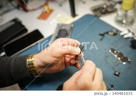 Hands hold a small part of an electronic device while tools and equipment are spread out on the workbench during the day. Hands hold a small part of an electronic device while tools and equipment are spread out on the workbench during the day. 140461035
