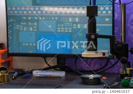 A microscope is positioned on a workstation next to a computer screen displaying a circuit board design. Tools are scattered. 140461037