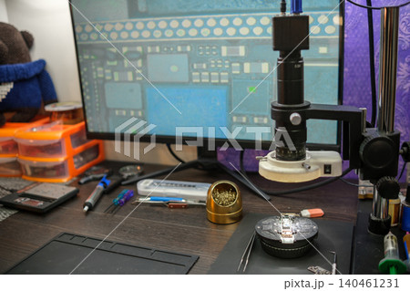 Tools and equipment lay on a desk next to a microscope and a monitor showing a circuit board, indicating work on electronic repairs. Tools and equipment lay on a desk next to a microscope and a monitor showing a circuit board, indicating work on electronic repairs. 140461231