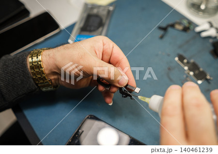 A person is repairing an electronic device by applying liquid to a small part while working on a blue surface. Tools and devices are nearby. 140461239