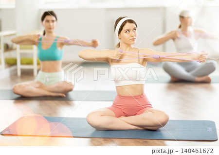 Sitting on mats in Pilates studio, young girls do arm stretches using an expander Sitting on mats in Pilates studio, young girls do arm stretches using an expander 140461672