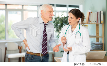 Senior man holds sore spot with his hand, complains about lower back pain to girl doctor 140462809
