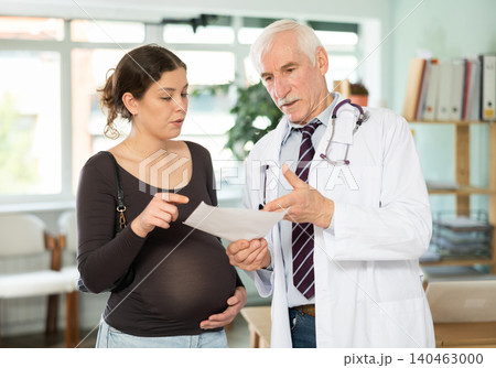 Senior man doctor and pregnant girl patient reviewing paper form, reading test results Senior man doctor and pregnant girl patient reviewing paper form, reading test results 140463000