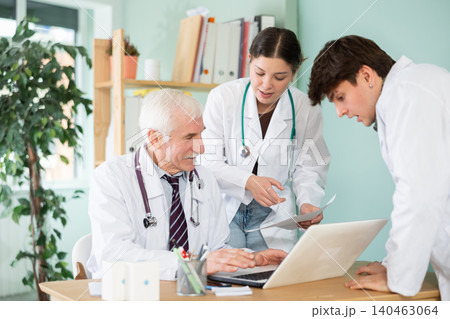 Professional elderly man doctor and two young male and female interns or nurses talking, working with laptop and checking patient papers in doctor office in clinic Professional elderly man doctor and two young male and female interns or nurses talking, working with laptop and checking patient papers in doctor office in clinic 140463064