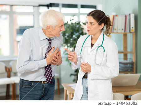 Elderly man patient complaining of pain to young woman doctor or nurse wearing white medical coat with stethoscope standing in office of medical center 140463082