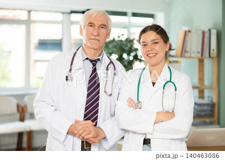 Two professional friendly medics in white medical coat standing with stethoscope and posing in clinic Two professional friendly medics in white medical coat standing with stethoscope and posing in clinic 140463086