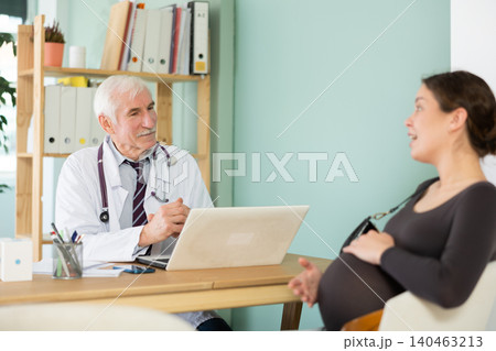 Pregnant woman expresses her complaints to an elderly male doctor sitting in front of laptop Pregnant woman expresses her complaints to an elderly male doctor sitting in front of laptop 140463213