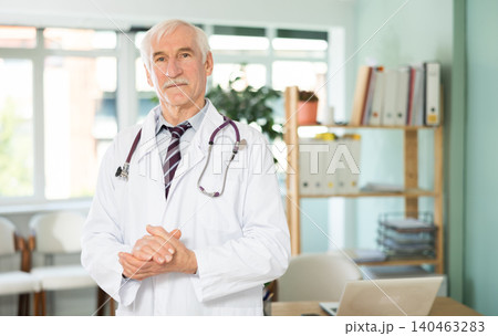 Portrait of friendly elderly man doctor wearing white uniform standing with stethoscope in clinic 140463283