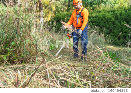 Landscaper trimming weeds with string trimmer in field, wearing safety gear and orange shirt 140464838