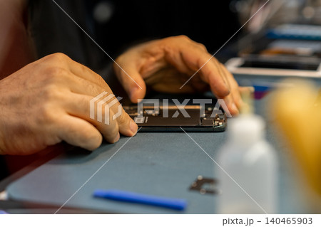 A skilled technician repairs a smartphone with focused attention on a workbench filled with tools and devices in a busy repair shop during the afternoon. A skilled technician repairs a smartphone with focused attention on a workbench filled with tools and devices in a busy repair shop during the afternoon. 140465903