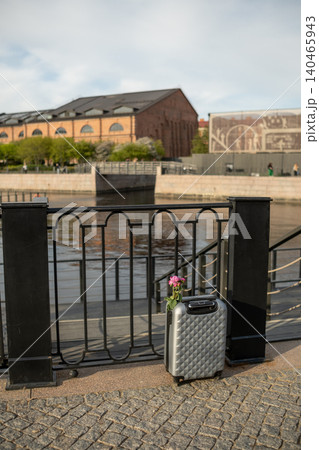 A silver suitcase adorned with flowers rests against a metallic railing by a tranquil waterfront in a lively urban area, inviting curiosity. A silver suitcase adorned with flowers rests against a metallic railing by a tranquil waterfront in a lively urban area, inviting curiosity. 140465943