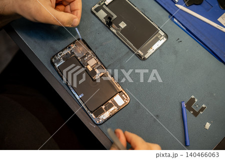 A technician focuses on repairing a smartphone, using tools to open the device and replace internal components on a work table filled with parts. A technician focuses on repairing a smartphone, using tools to open the device and replace internal components on a work table filled with parts. 140466063