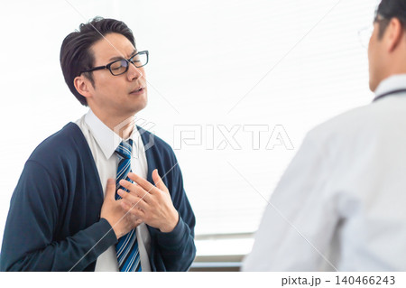病院の診察室で医師に胸の苦しみや痛みを訴えるネクタイ姿の男性患者 140466243