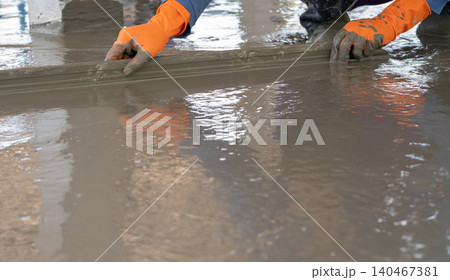 Mason worker working with trowel to level cement floor at construction site. A skilled construction worker spreading wet concrete to level wet concrete on a site floor. Foundation and flooring tasks. Mason worker working with trowel to level cement floor at construction site. A skilled construction worker spreading wet concrete to level wet concrete on a site floor. Foundation and flooring tasks. 140467381