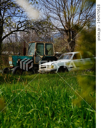 Old abandoned rusty tractor and white vintage sedan car in a green field  140470628
