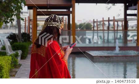 Woman wearing traditional red jalabiya using smartphone by canal Woman wearing traditional red jalabiya using smartphone by canal 140473510