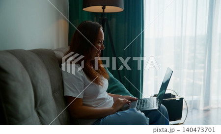 Woman working on laptop while sitting on couch by window Woman working on laptop while sitting on couch by window 140473701