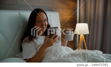 Woman in bed holding coffee cup browsing smartphone in morning relaxation Woman in bed holding coffee cup browsing smartphone in morning relaxation 140473761