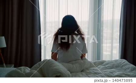 Woman sitting on bed gazing through window at morning light Woman sitting on bed gazing through window at morning light 140473895