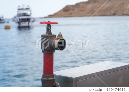 Red fire hydrant valve positioned on a dock with boats visible in the background and calm water reflecting the nearby shoreline Red fire hydrant valve positioned on a dock with boats visible in the background and calm water reflecting the nearby shoreline 140474611