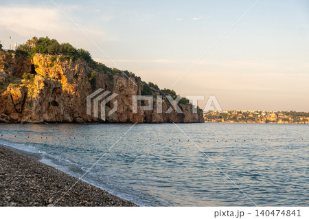 Cliffs rise above a calm sea as the sun sets, casting warm hues on the water, while pebbles line the beach in a serene coastal atmosphere. Cliffs rise above a calm sea as the sun sets, casting warm hues on the water, while pebbles line the beach in a serene coastal atmosphere. 140474841