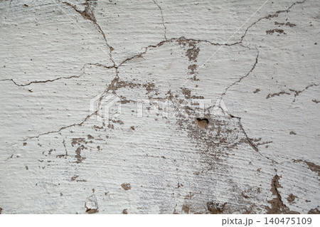 Surface of a cracked white wall shows uneven texture and signs of age, with faint marks and a small hole, creating an impression of neglect. 140475109