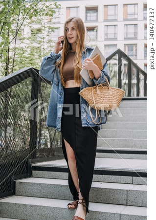 Young woman talks on smartphone while walking down city stairs holding laptop. Working on the go, independent creative, flexible remote job, Modern content creator. 140477137