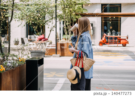 Woman standing outdoors sips beverage while holding smartphone and carrying woven tote bag. Sustainable living, urban lifestyle, eco friendly choices. Woman standing outdoors sips beverage while holding smartphone and carrying woven tote bag. Sustainable living, urban lifestyle, eco friendly choices. 140477273