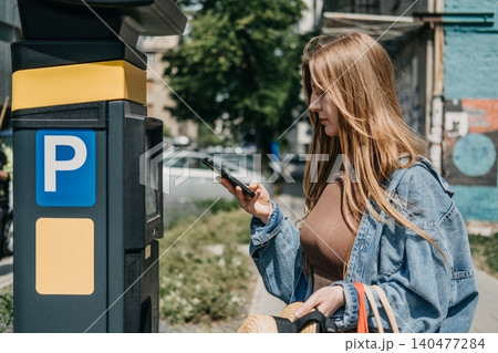 Woman looks at smartphone while standing next to city parking meter terminal. Ticketless parking technology, connected urban infrastructure, municipal fintech application Woman looks at smartphone while standing next to city parking meter terminal. Ticketless parking technology, connected urban infrastructure, municipal fintech application 140477284