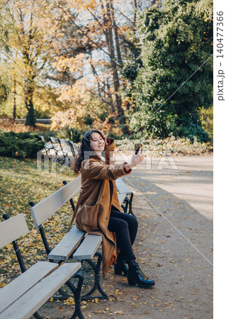 Woman sits on bench in public park while taking selfie or recording video with smartphone. User generated content UGC production, mobile photography in nature, social media marketing imagery. Woman sits on bench in public park while taking selfie or recording video with smartphone. User generated content UGC production, mobile photography in nature, social media marketing imagery. 140477366
