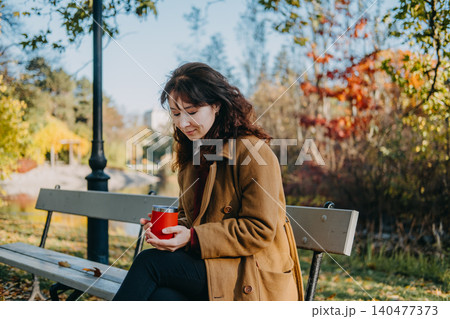 Woman sits on park bench holding insulated mug during autumn day. Mental health break, nature therapy, outdoor routine. 140477373