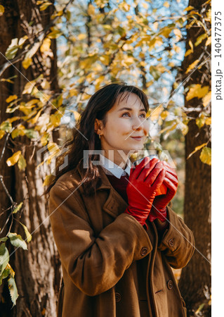 Woman stands in woods holding beverage container with both hands while looking upward. Mindfulness outdoor breathing exercise, positive thinking mental health, peaceful nature walk reflection. Woman stands in woods holding beverage container with both hands while looking upward. Mindfulness outdoor breathing exercise, positive thinking mental health, peaceful nature walk reflection. 140477375