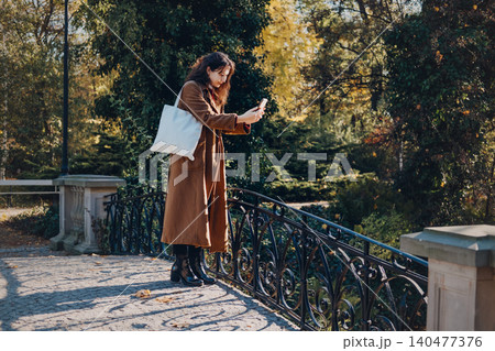 Woman stands on pedestrian bridge holding smartphone to take photograph. Taking photo with smartphone camera, outdoor content creator lifestyle, millennial blogger capturing moment. Woman stands on pedestrian bridge holding smartphone to take photograph. Taking photo with smartphone camera, outdoor content creator lifestyle, millennial blogger capturing moment. 140477376