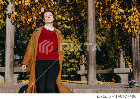 Woman smiles while walking outdoors near trees with autumn foliage. Expressing pure happiness, positive mental health, mindfulness in daily life, optimism and wellbeing. Woman smiles while walking outdoors near trees with autumn foliage. Expressing pure happiness, positive mental health, mindfulness in daily life, optimism and wellbeing. 140477400