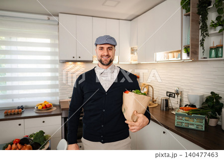 Man smiling holding paper bag with fresh groceries in kitchen Man smiling holding paper bag with fresh groceries in kitchen 140477463