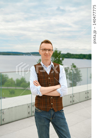 Middle-aged man poses with arms crossed on observation desk by water with cloudy sky and green trees in background near modern building Middle-aged man poses with arms crossed on observation desk by water with cloudy sky and green trees in background near modern building 140477775