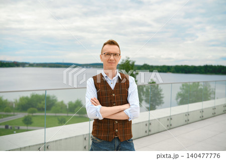 Middle-aged man stands on balcony of observation deck above lake in park with his arms crossed over his chest Middle-aged man stands on balcony of observation deck above lake in park with his arms crossed over his chest 140477776