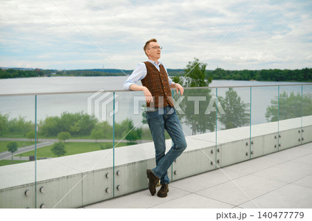Middle-aged man in vest and jeans leans against glass railing with river in background 140477779
