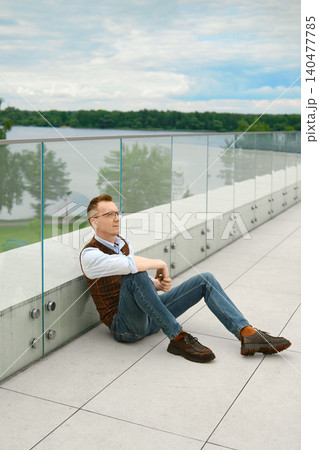 Relaxed middle-aged man sitting by lake on modern terrace during cloudy weather Relaxed middle-aged man sitting by lake on modern terrace during cloudy weather 140477785